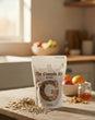 Bag of 'The Granola Kid' granola on a wooden table with fruits and a glass in the background